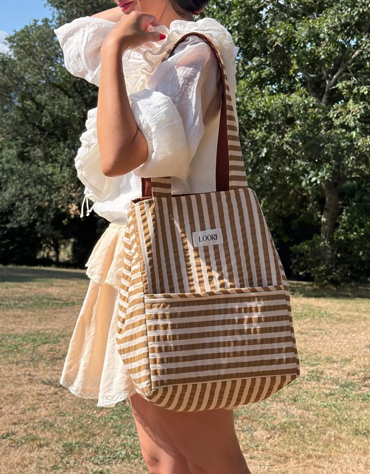 Mujer llevando al hombro el bolso LOÖRI de rayas camel en un prado